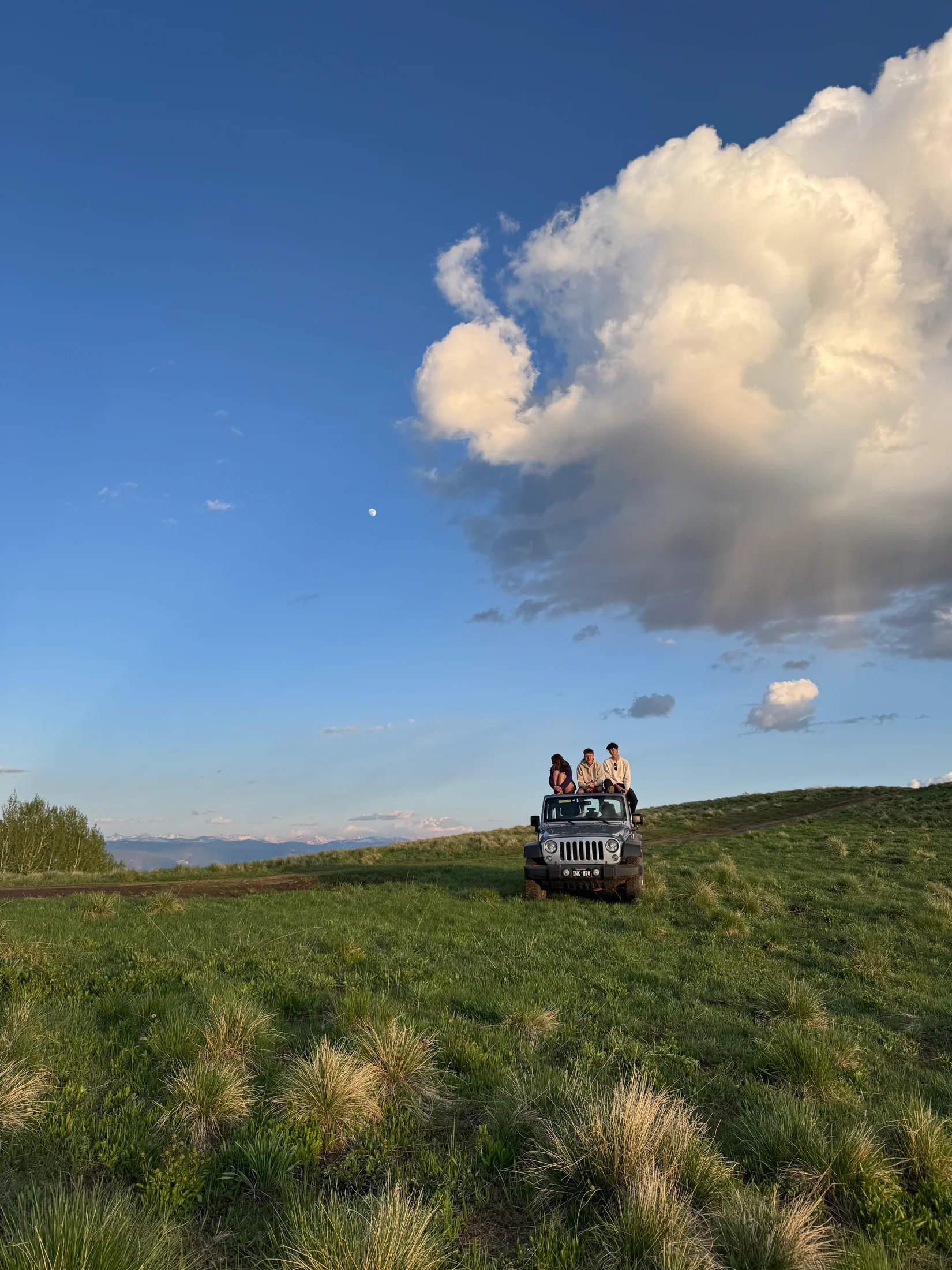 Judah's Jeep parked on a high green ridge, friends on the roof beneath a towering evening sky